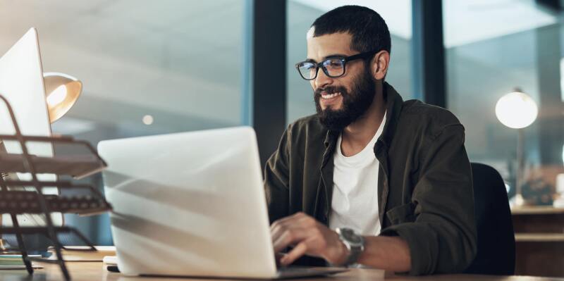 Man in cardigan and T-shirt smiling while he works on laptop on table