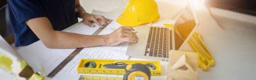 Civil engineer working with blueprints on a desk in front of a laptop surrounded by measuring tools and safety helmet