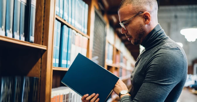 Male student wearing glasse in a library, reviewing a book of interest.