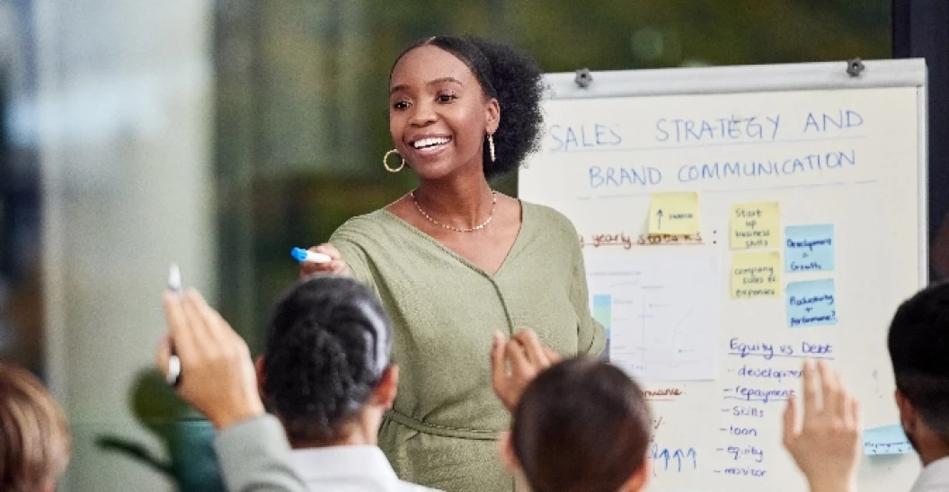 Woman at white board in front of room of colleagues raising hands