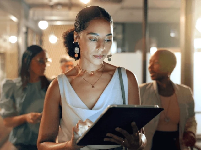A woman efficiently managing tasks on her iPad, with coworkers engaged in communication behind her.